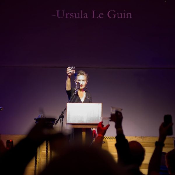 Woman (Annetta Black) stands on stage and raises a glass in front of a crowd